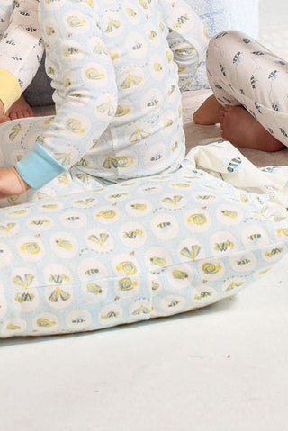 Three children in light pajamas with bee and flower prints sit on the floor. In the foreground is a Buggin’ Out Children’s Sleeping Pillow Cover, adding a playful touch to this cozy scene. Their faces are not visible.