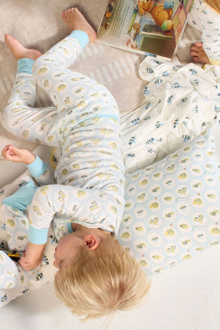 A young child with blonde hair in patterned pajamas lies on a Buggin’ Out Children’s Sleeping Pillow Cover, next to a blanket and an open picture book, on a light-colored surface.