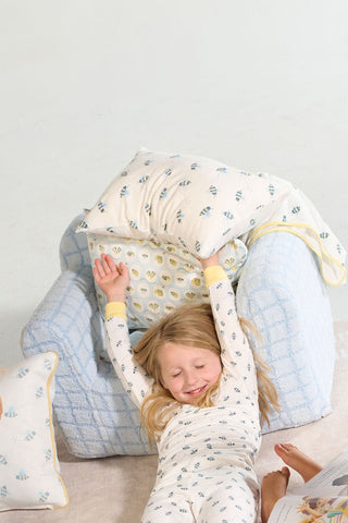 A young child in patterned pajamas stretches and smiles while reclining on a cozy blue armchair with a Buzzin’ Around Children’s Sleeping Pillow Cover, creating a comfortable and relaxed scene.
