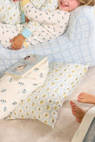 Two kids in patterned pajamas sit on a soft blue blanket with pillows, including the Buggin’ Out Children’s Sleeping Pillow Cover, and an open book. Only parts of their faces and legs are visible.