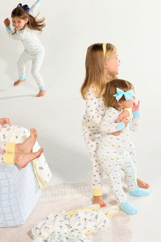 Three young children play together; one hugs a younger child wearing the Buggin’ Out Dream & Play Zipper Romper with a hand-drawn bug print, while another jumps in the background. A blanket and feet are visible on the couch.