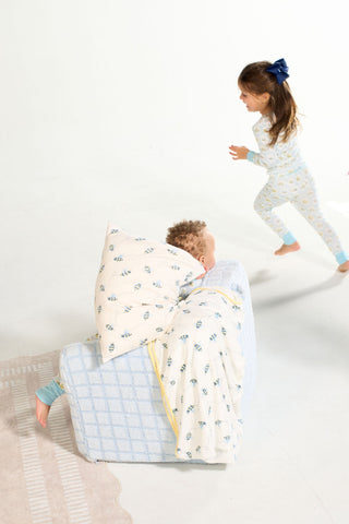 A young child lounges on a chair with the Buzzin’ Around Children’s Blanket, while another child in pajamas with a blue bow runs by on a light-colored floor.