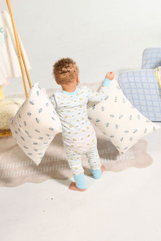 A toddler in pajamas stands on a rug holding two large pillows with Buzzin’ Around Children’s Sleeping Pillow Cover, near a light blanket and hanging clothes in a bright, cozy room.