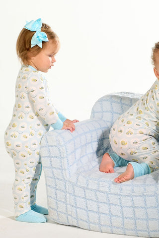 Two toddlers in matching light blue Buggin’ Out Dream & Play Zipper Rompers and socks play on a fuzzy blue-checkered couch—one stands by the armrest, the other kneels with their back turned.