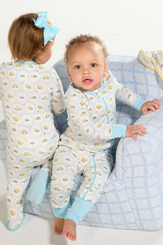 Two toddlers in Buggin’ Out Dream & Play Zipper Romper pajamas with blue trim and a bug print play on a cozy chair. One faces forward; the other, wearing a blue bow, is turned away.