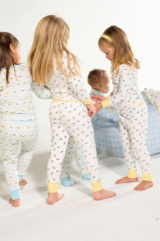 Four kids wear the Buzzin’ Around Dream & Play Two Piece Set as they stand holding hands, while a toddler sits on a blue plaid cushion in the cozy, softly lit room.