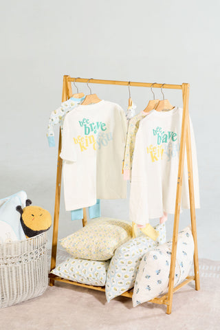 Two white Bee You Long Sleeve Children’s Tees hanging on a wooden rack, each printed with the words bee brave, bee kind, bee you in stacked, colorful lettering.