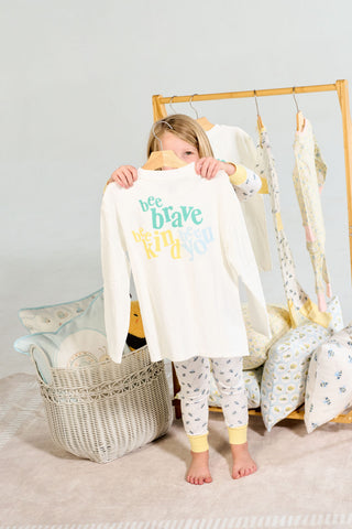A child holds up a white Bee You Long Sleeve Children’s Tee featuring a bee graphic and the colorful text bee brave, bee kind, bee you.