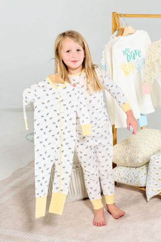 A young girl in bee-patterned pajamas holds up a Buzzin’ Around Dream & Play Zipper Romper. Behind her, a rack displays more children’s clothes and pajamas in a bright, cheerful setting.