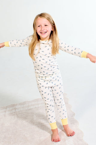 Smiling child with long blonde hair stands barefoot on a light rug, wearing the Buzzin’ Around Dream & Play Two Piece Set—soft cotton modal pajamas with a small blue pattern and yellow cuffs, arms outstretched playfully.