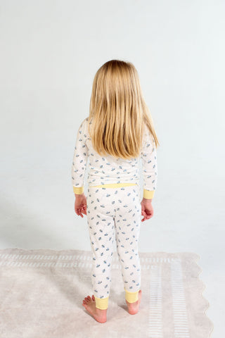 A young child with long blonde hair stands barefoot on a rug, facing away, wearing Buzzin’ Around Dream & Play Two Piece Set—white cotton modal pajamas with a small blue pattern and yellow cuffs.