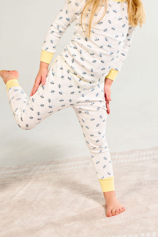 A child in the Buzzin’ Around Dream & Play Two Piece Set stands on one leg, holding the other behind them, on a light-colored carpet. The white cotton modal pajamas have a blue pattern and yellow cuffs.