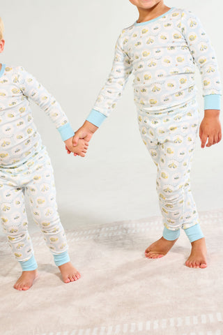Two children in matching Buggin’ Out Dream & Play Two Piece Set cotton modal pajamas stand barefoot on a rug, holding hands. Their cozy baby sleepwear has blue cuffs and yellow patterns, faces out of frame.