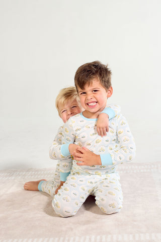 Two young kids in Buggin’ Out Dream & Play Two Piece Set sit on a soft rug. The front child smiles widely as the one behind hugs him, both playful with light brown hair and a plain, light background.