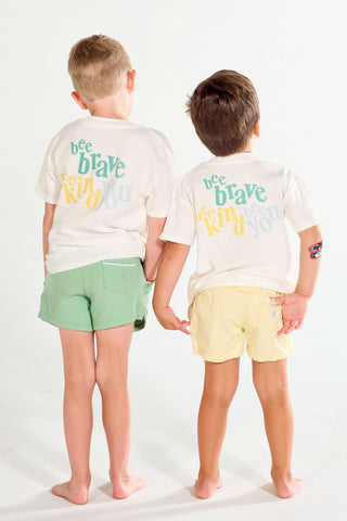 Two young boys seen from behind wearing matching Bee You Short Sleeve Children’s Tee shirts with colorful lettering, one in green shorts and the other in yellow shorts.