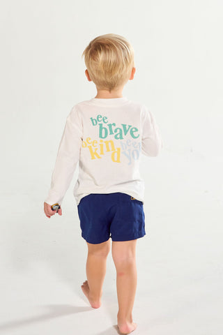 A toddler seen from behind wearing a Bee You Long Sleeve Children’s Tee with bee brave, bee kind, bee you printed on the back in colorful lettering, paired with blue shorts.