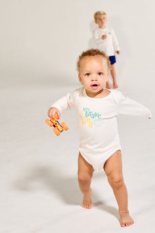 A toddler in a white Bee You Long Sleeve Onesie holding a small toy truck, with a young boy standing in the background.