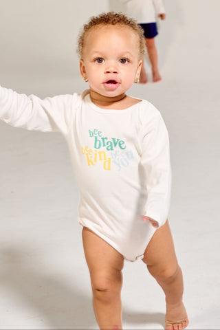 A toddler wearing a white Bee You Long Sleeve Onesie stands in the foreground, with another child blurred in the background.