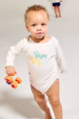 A toddler wearing a white Bee You Long Sleeve Onesie printed with the words bee brave, bee kind, bee you holds a toy monster truck while another child stands in the background.