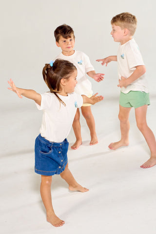 Three children wearing white hand-drawn Bee You Short Sleeve Children’s Tee shirts stand in a white studio, with one child in the foreground with arms outstretched and two children standing behind.