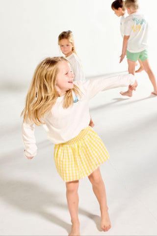 A girl wearing a Bee You Long Sleeve Children’s Tee and a yellow gingham skirt dances in a bright studio, with two other children in the background.