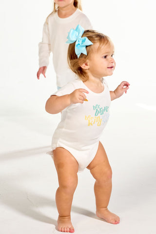 A toddler wearing a white Bee You Short Sleeve Onesie with bee brave text and a large light blue hair bow, with a slightly older child walking behind her.