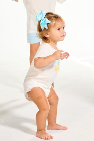 A toddler wearing a white short-sleeve Bee You Short Sleeve Onesie and a light blue hair bow stands in front of a dancing child.