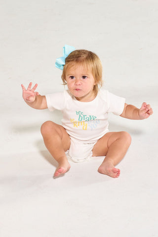 A toddler wearing a white Bee You Short Sleeve Onesie sits on a white floor while another child stands nearby holding a toy.