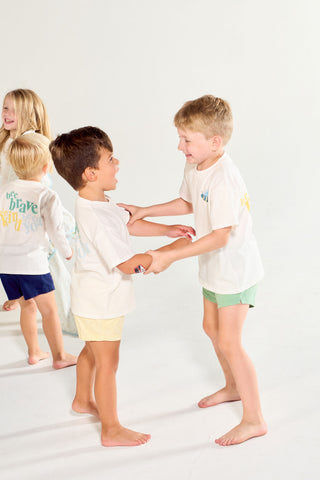 Two young boys wearing hand-drawn Bee You Short Sleeve Children’s Tee shirts hold hands and laugh, while another child stands in the background.