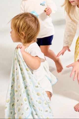 A toddler walks barefoot holding the Buggin’ Out Children’s Blanket, while another child reaches out, their feet visible on a light floor.
