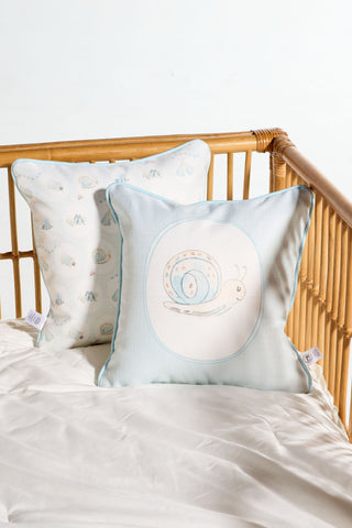 Two light blue Buggin’ Out Children’s Pillows on a light wooden crib: one with a snail illustration, the other with a repeating seashell pattern, both on a crisp white sheet.
