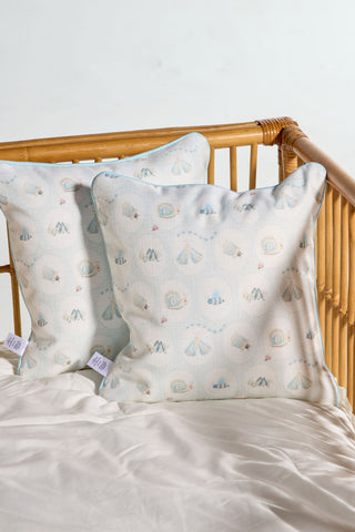 Two Buggin’ Out Children’s Pillows with light blue and tan seashell patterns rest against crib slats on a cream-colored sheet.