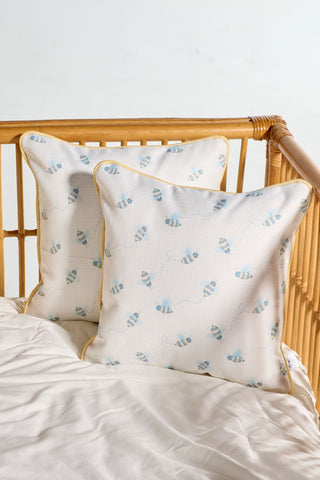 Two Buzzin’ Around Children’s Pillows, featuring light blue bee patterns and yellow piping, rest in a wicker crib beside a white sheet, creating a cozy, airy vibe.