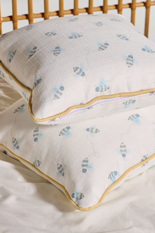 Two Buzzin’ Around Children’s Pillows with blue and yellow bee prints and yellow piping are stacked on a white bed, adding charm and a playful touch to kids’ rooms beneath a wooden headboard.