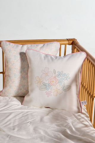 Two Precious Petals Children’s Pillows with pink piping rest against the side of a light wooden crib with a white sheet. The pillow in front features embroidered flowers, while the one in back displays an allover floral pattern. Both are machine washable for easy care.