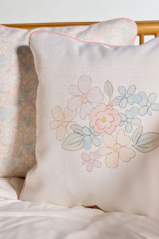 The Precious Petals Children’s Pillow, featuring pastel flower embroidery, sits in front of a matching floral-patterned pillow on a light bedding set. Thoughtfully made in the USA, this accent is both stylish and machine washable for easy care.