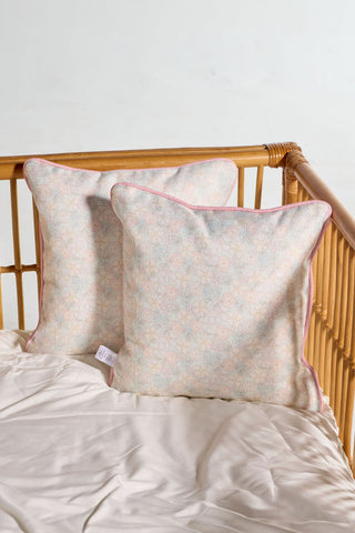 Two light-colored Precious Petals Children’s Pillows, featuring a floral pattern with pink edging, rest against the side of a wooden crib. The crib is dressed in beige bedding and set against a light, neutral background. These pillows are machine washable for easy care.