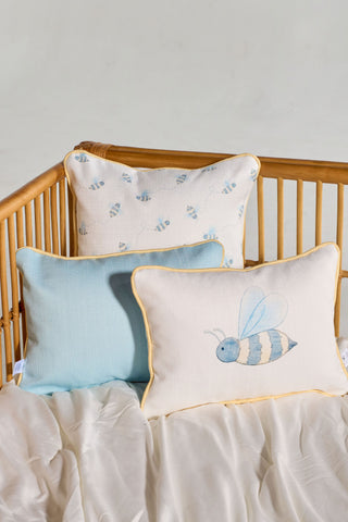 The Bee You Children’s Lumbar Pillow, showcasing a large bee illustration, sits beautifully arranged alongside a bee-patterned pillow and a solid light blue pillow on a white bed with a light wooden headboard. Each machine washable pillow is thoughtfully designed to offer both comfort and style.