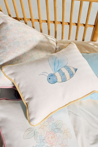 The Bee You Children’s Lumbar Pillow, featuring a blue and white bee illustration, rests on a wicker chair among other pastel-hued pillows with floral designs. This charming accent offers double-sided prints for added versatility and is crafted to be machine washable, ensuring easy care.