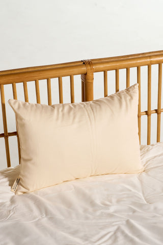 An Organic Children’s Sleeping Pillow in a cream color rests on a neatly made bed with white sheets and a light wooden slatted headboard in the background.