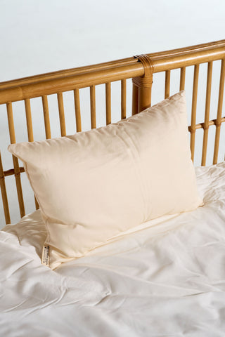 An Organic Children’s Sleeping Pillow in a cream color rests on a white bed with a wooden slatted headboard.