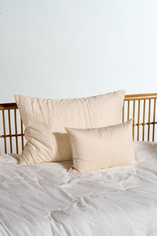 Two cream-colored pillows, one large and one small, rest against a light wooden headboard on a bed with white sheets—the smaller cushion is the Organic Children’s Sleeping Pillow, designed to be a gentle, hypoallergenic kids pillow for your child’s restful sleep.