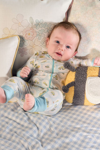 A baby wearing the Buggin’ Out Dream & Play Zipper Romper with a hand-drawn bug print smiles on a bed with floral and checkered pillows, holding a plush bee toy.