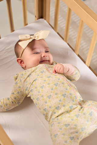 A baby in the Precious Petals Dream & Play Zipper Romper and a cream-colored bow headband smiles while lying on a white sheet in a wooden crib.