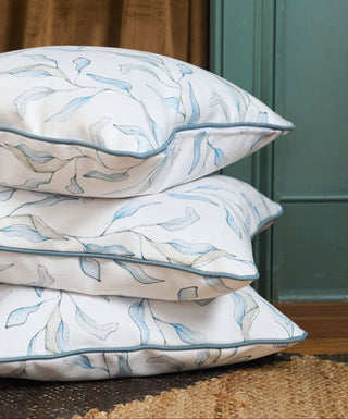 Three stacked decorative throw pillows with a blue and beige botanical leaf pattern on a white background, featuring light blue piping, set against a background of brown curtains and a teal door.