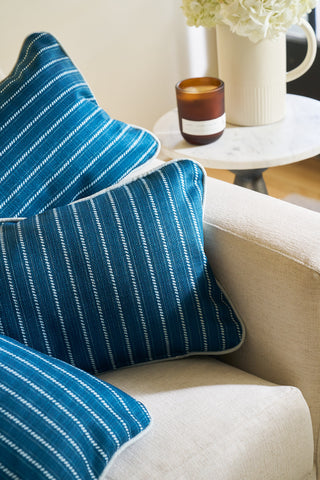 The Courtyard Stripes Extended Lumbar Pillow from the Core Collection sits on a light sofa beside a small round table with a brown candle and white vase filled with Broad Street Blooms.