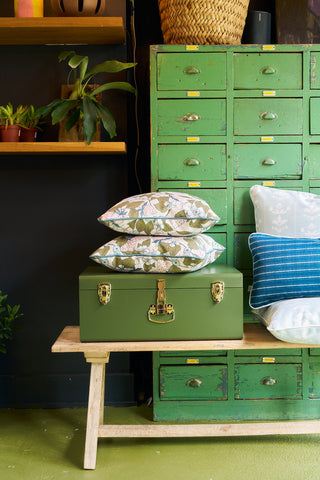 A green metal trunk with the Fruit Fields Pillow rests on a wooden bench by vintage green drawers, surrounded by potted plants and a woven basket for cozy, rustic charm in eclectic garden decor.