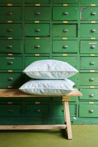 Two Garden Party Pillow covers in light blue floral from the Core Collection are stacked on a wooden bench in front of a vintage green cabinet with small drawers and yellow labels.