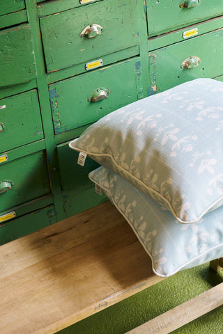 Two Garden Party Pillows in light blue with white botanical patterns, from the Core Collection, are stacked on a wooden bench set before vintage green drawers featuring metal handles and label slots.