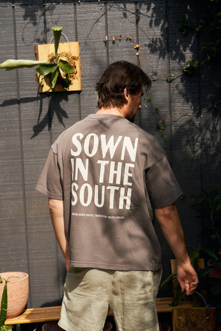 A person stands outdoors with their back to the camera, wearing a gray Sown in the South Short Sleeve Tee featuring "SOWN IN THE SOUTH" in bold white letters. Plants and a gray wooden wall are visible in the background.